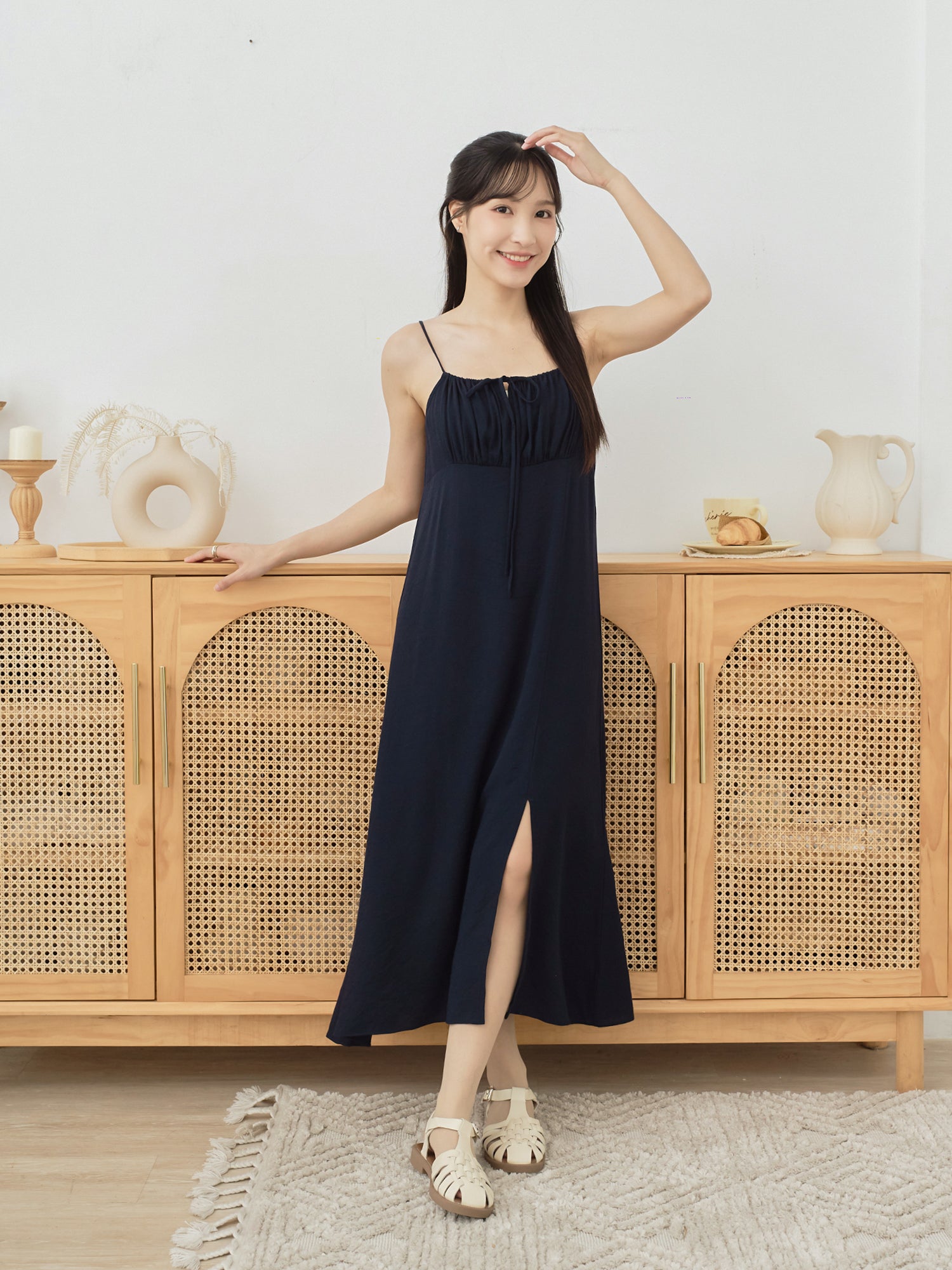 A woman modeling a navy blue sleeveless maxi dress with thin straps, a gathered neckline, and a front slit, standing in front of a light wood cabinet with woven doors in a bright indoor setting.