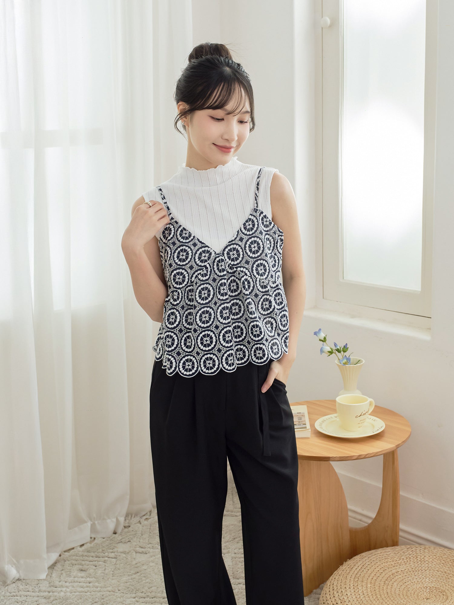 A woman stands indoors wearing a sleeveless black-and-white geometric patterned camisole over a white mock-neck top, paired with black wide-leg pants. She is posed beside a small wooden side table with a cup and saucer and a small vase of flowers in a bright room with white curtains and a window.