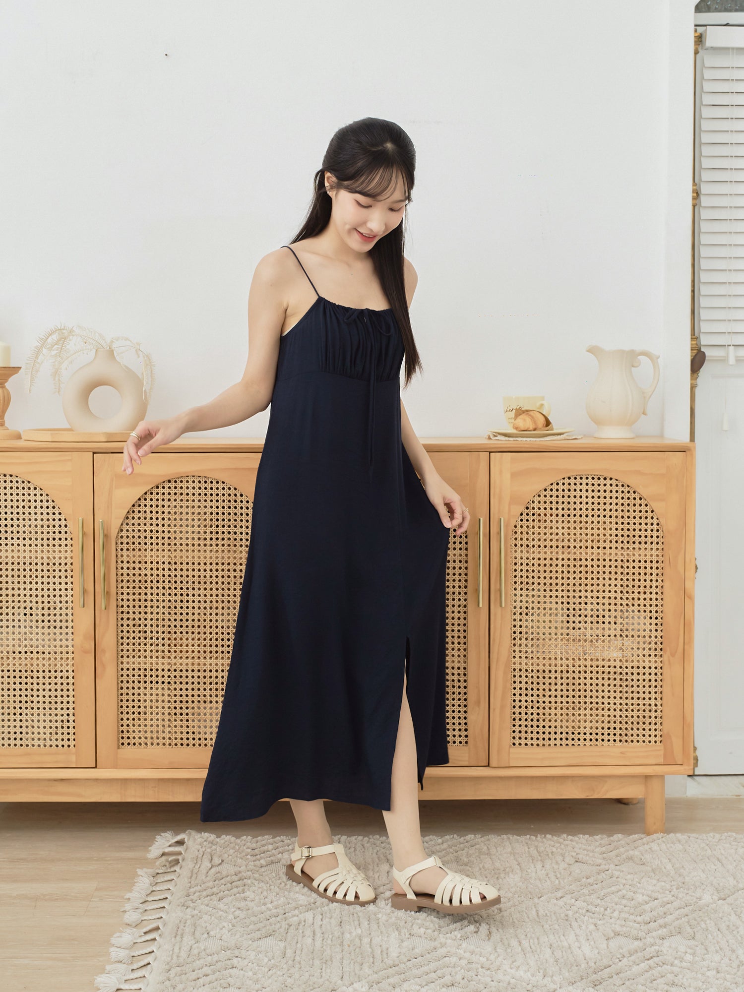 A woman modeling a dark navy midi-length spaghetti-strap dress with a gathered neckline and a front slit, standing in front of a light wood cabinet with woven cane doors.