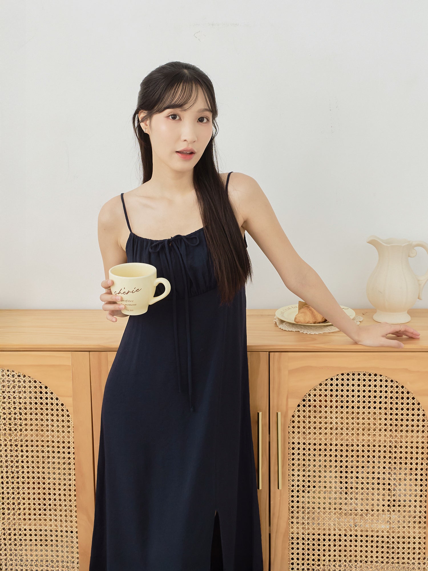 A woman wearing a dark navy blue spaghetti-strap maxi dress stands in a bright room, holding a cream-colored ceramic mug. She is positioned in front of a light wood cabinet with woven cane panels and a white wall behind her.